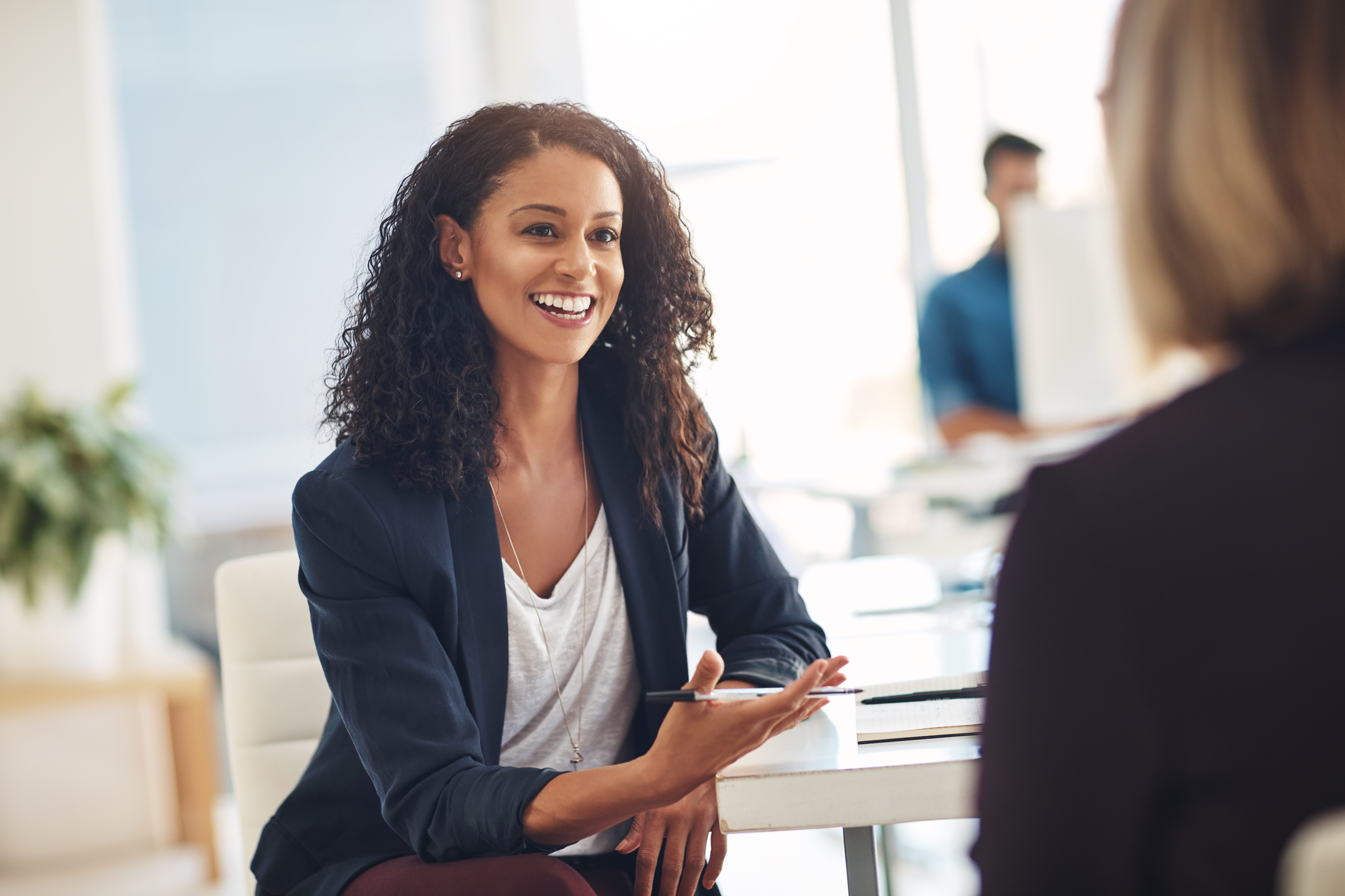 Two women sitting at a desk in business wear with the perspective being over one of their shoulders
